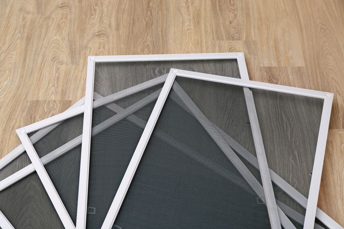 Several rectangular window screens with white frames are stacked on a light wood floor.