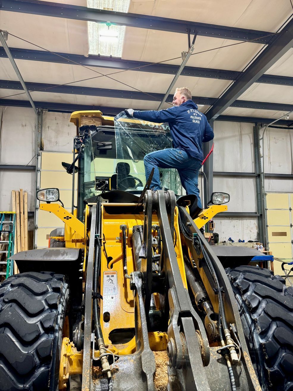 Man standing on a yellow front-end loader inside a warehouse, reaching toward the windshield.