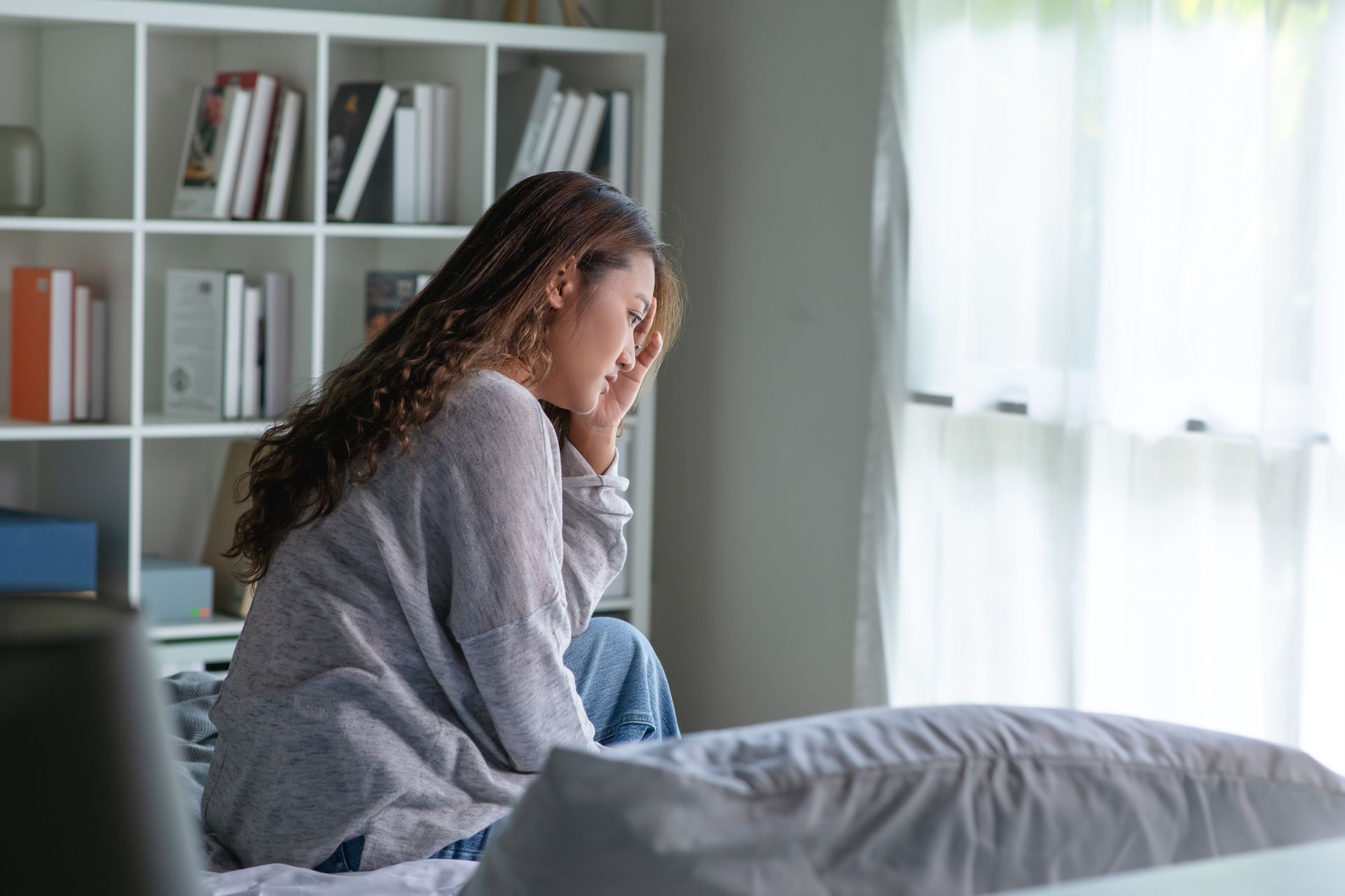 Woman sitting on a bed, looking distressed. She holds her head near a window and bookshelf.