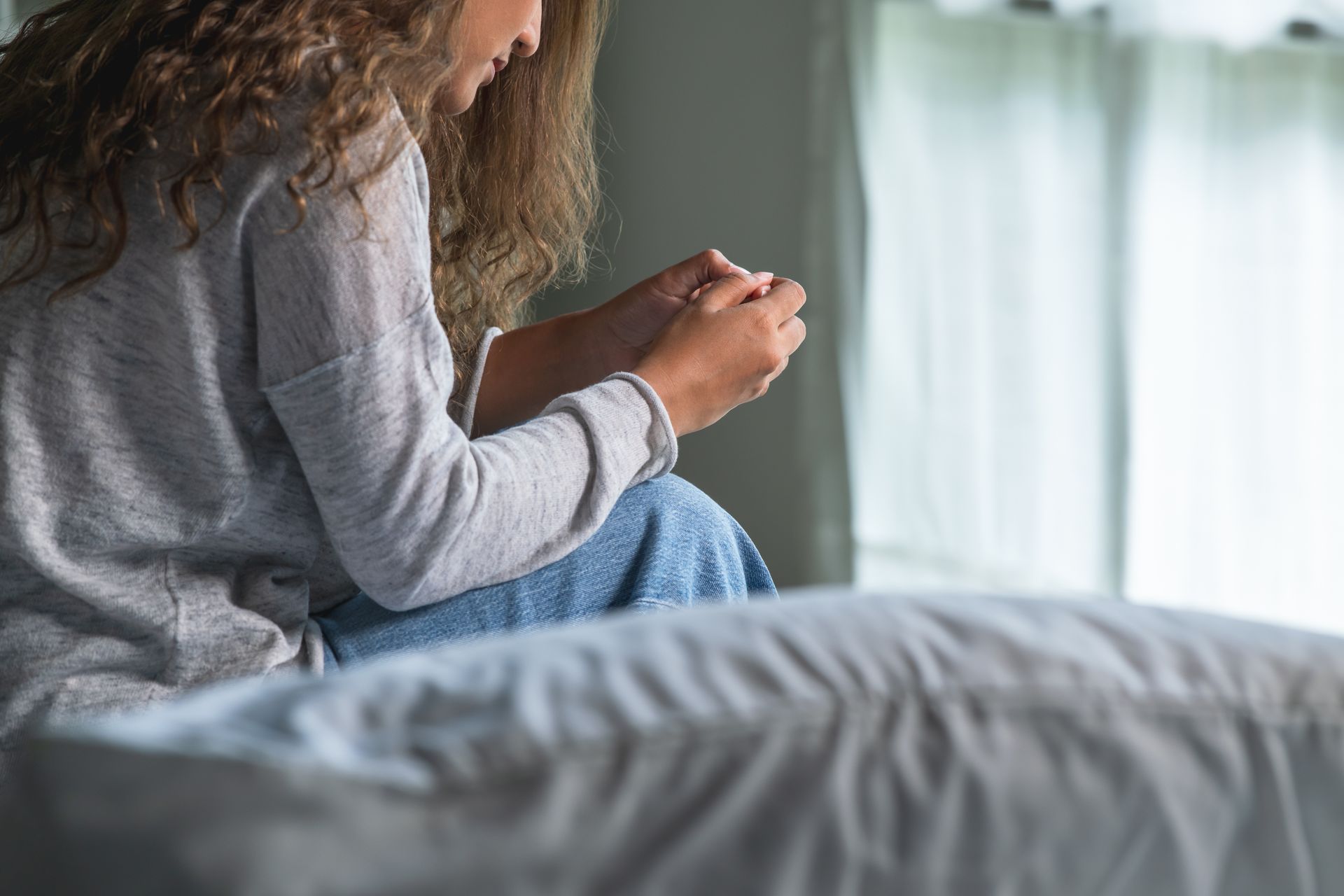 Woman sitting on bed, head down, hands clasped. Natural light from window.