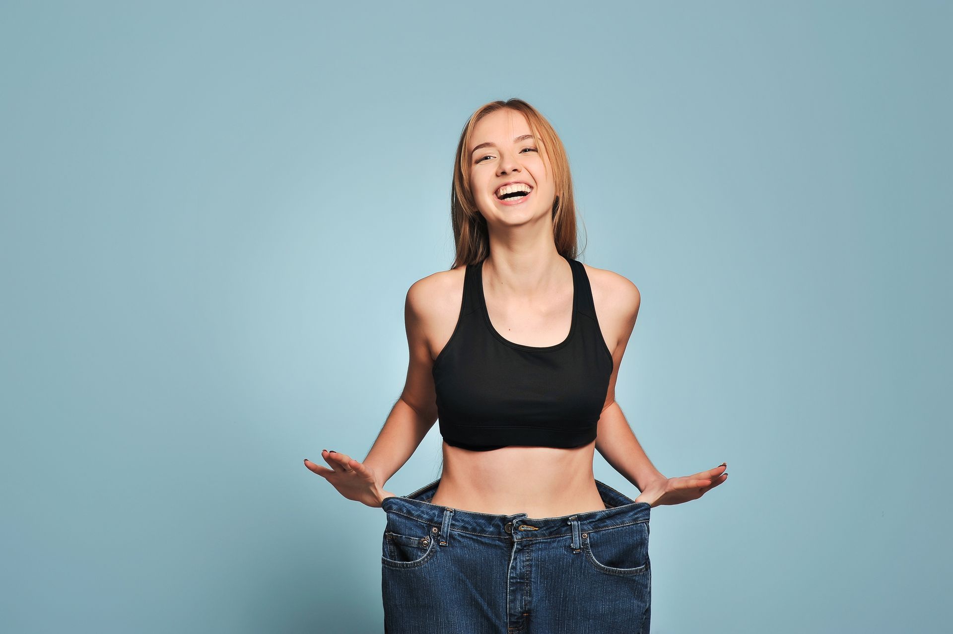 Woman wearing oversized jeans, smiling, holding out sides, showing weight loss. Light blue background. Woman wearing oversized jeans, smiling, holding out sides, showing weight loss. Light blue background.