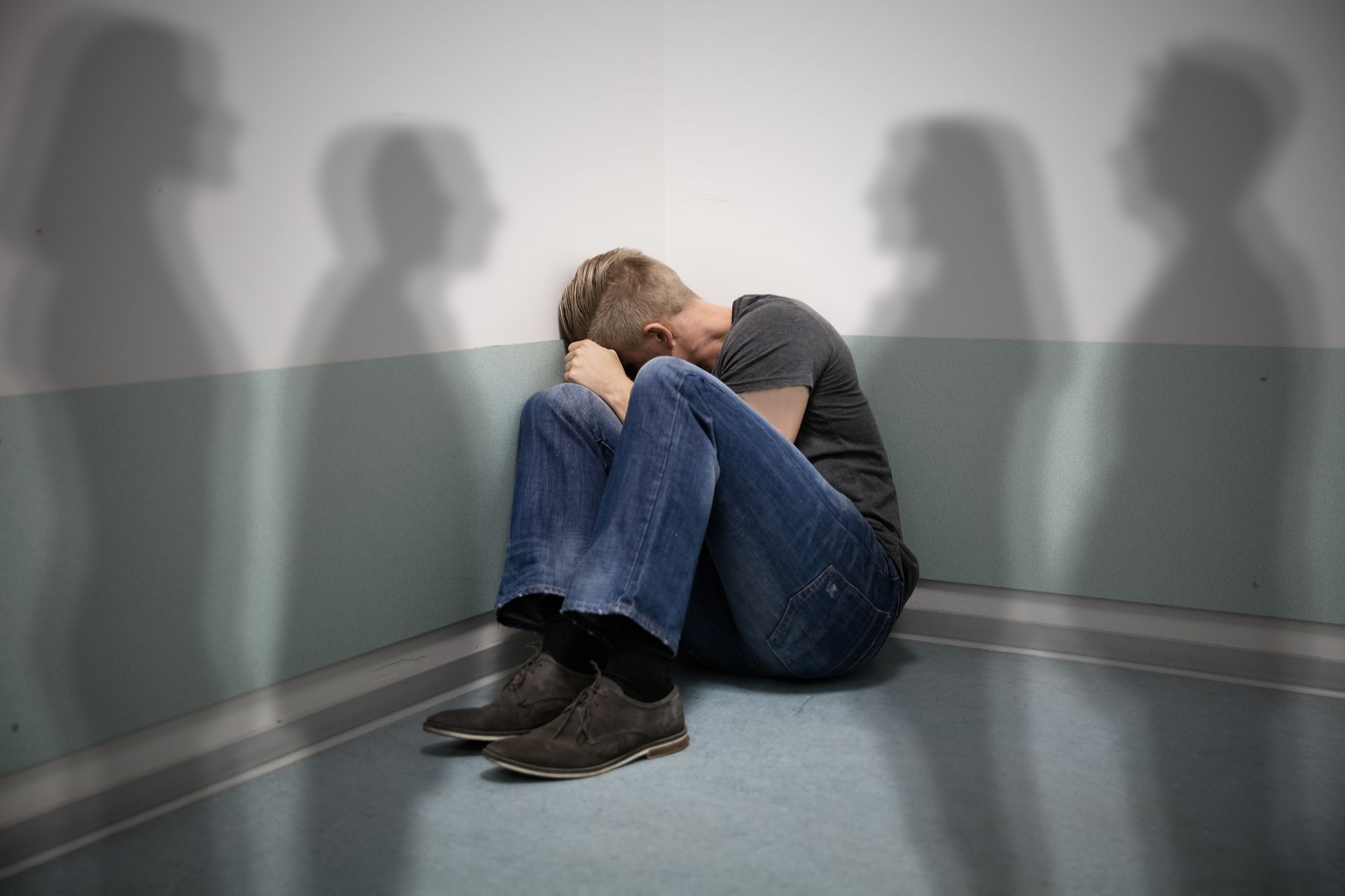 Person in jeans and a t-shirt sitting in a corner, head in hands. Shadows of people are cast on the wall. Person in jeans and a t-shirt sitting in a corner, head in hands. Shadows of people are cast on the wall.