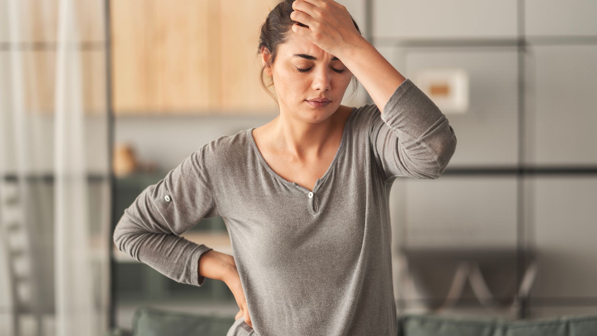 Woman holding her head and back, appearing to be in pain, indoors. Woman holding her head and back, appearing to be in pain, indoors.