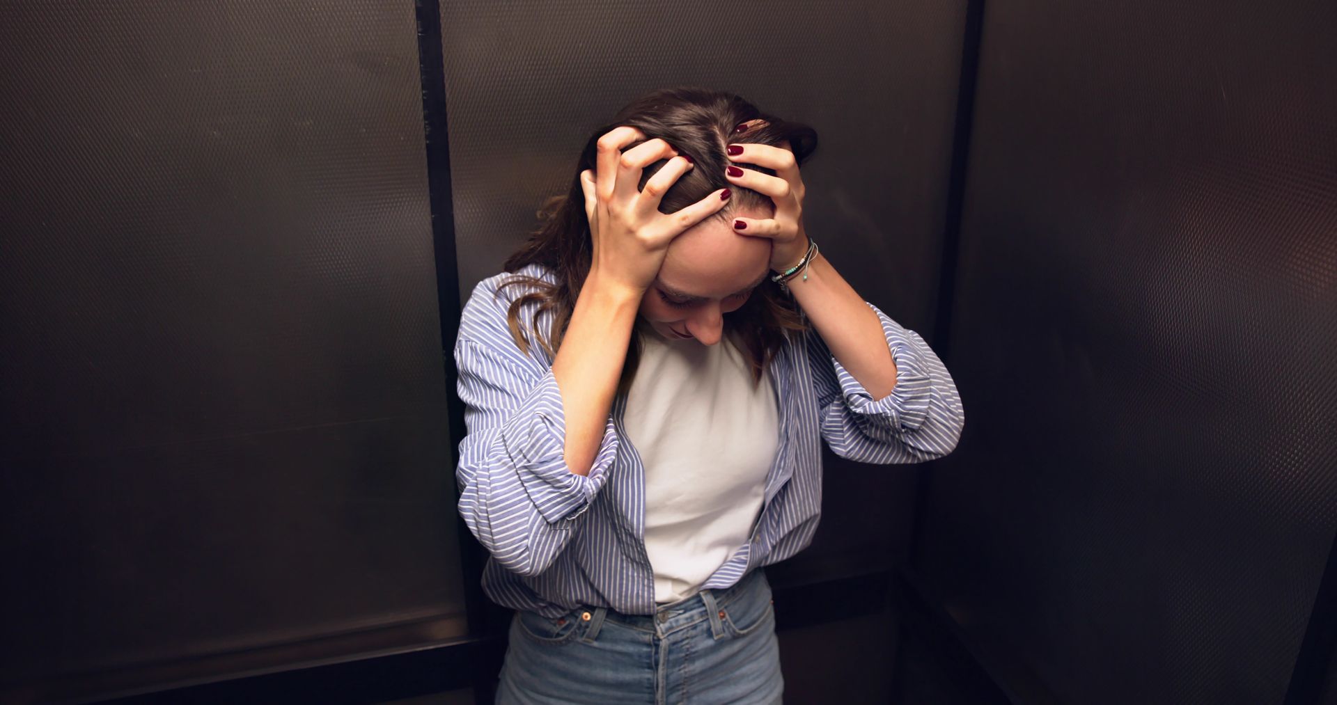 Woman in a striped shirt, distressed, clutches her head in a dark elevator.