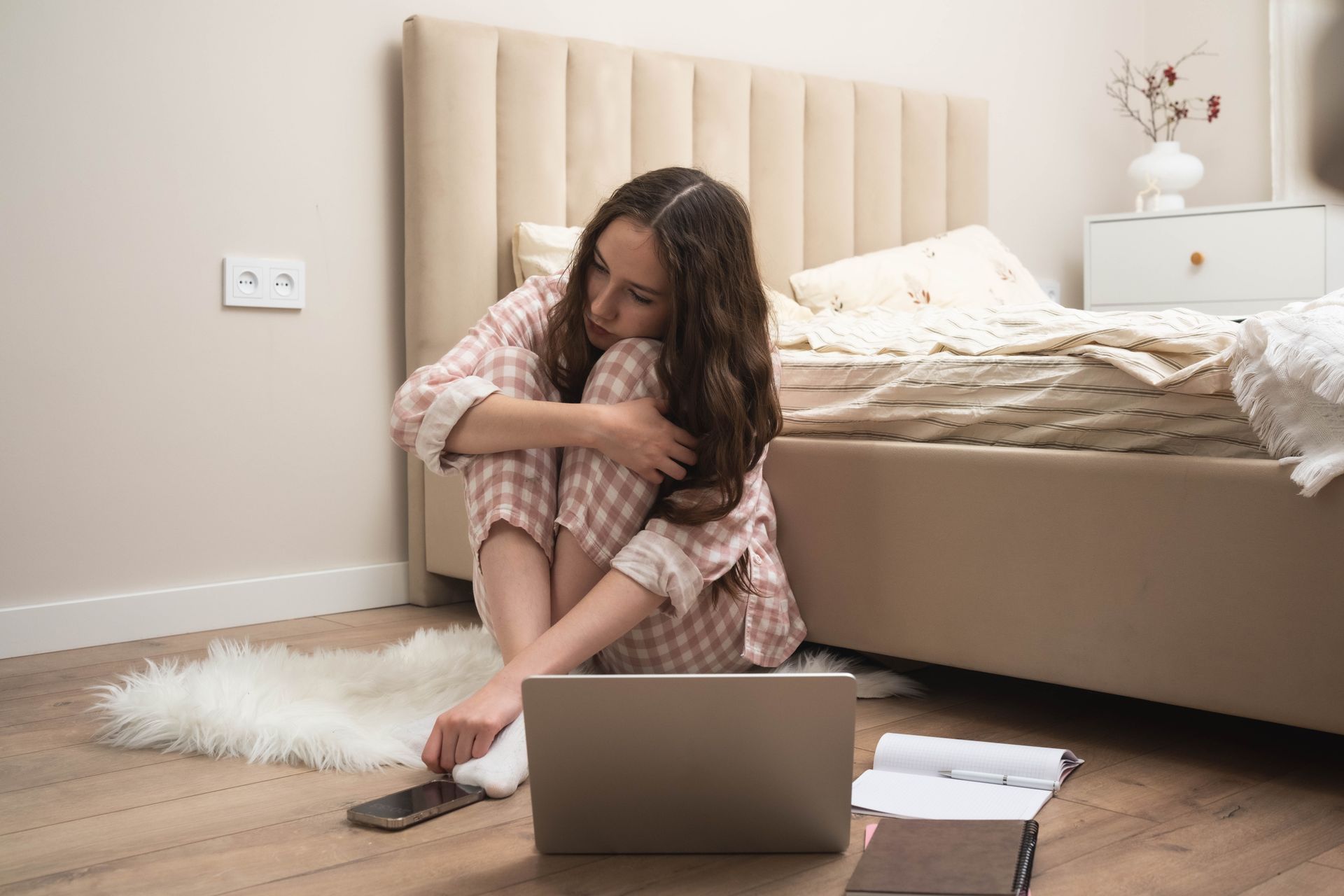 Woman in pajamas curled up on floor by laptop and bed, looking down, next to a rug. Woman in pajamas curled up on floor by laptop and bed, looking down, next to a rug.