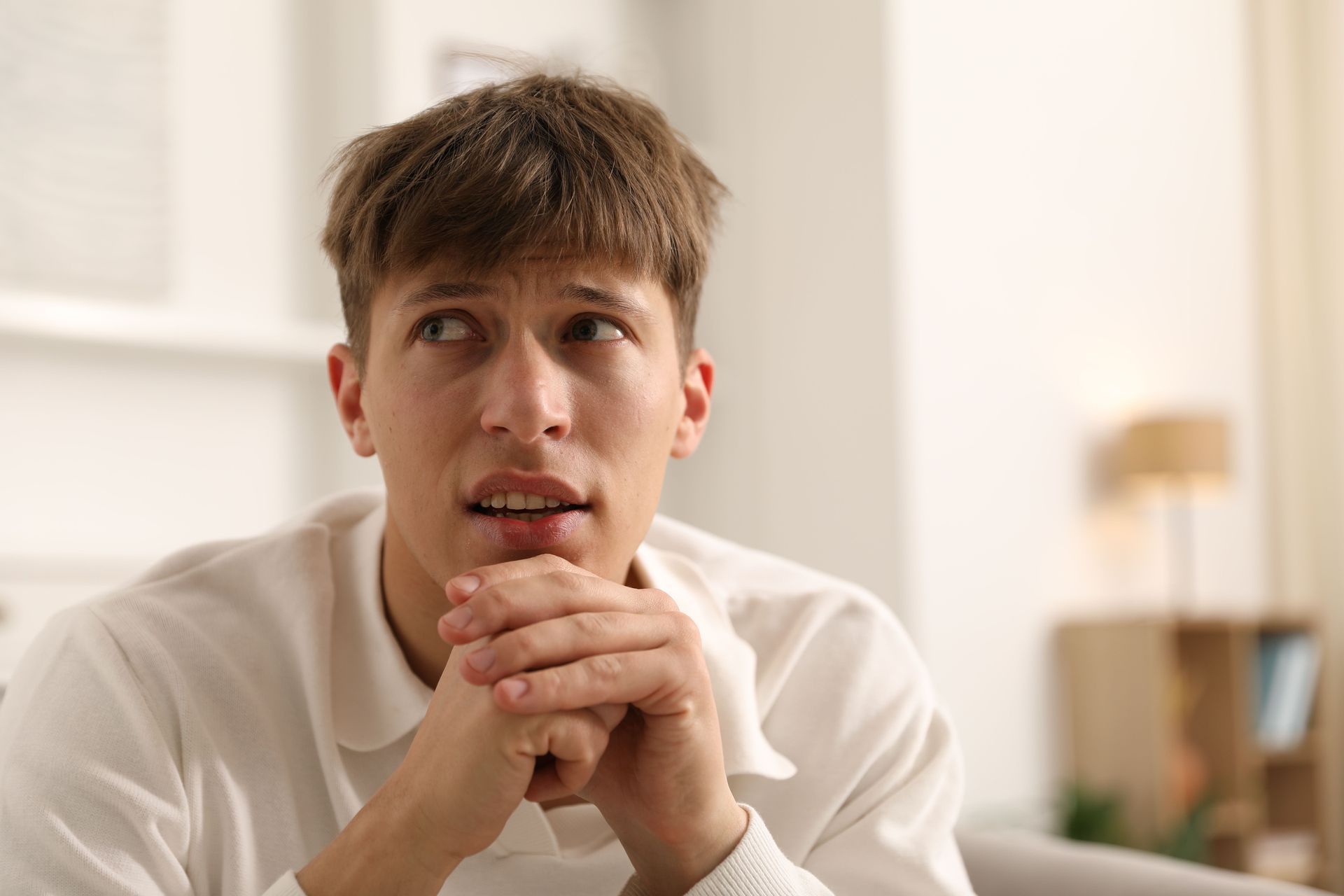 Young person with hands clasped, looking away with a concerned expression, indoors. Young person with hands clasped, looking away with a concerned expression, indoors.