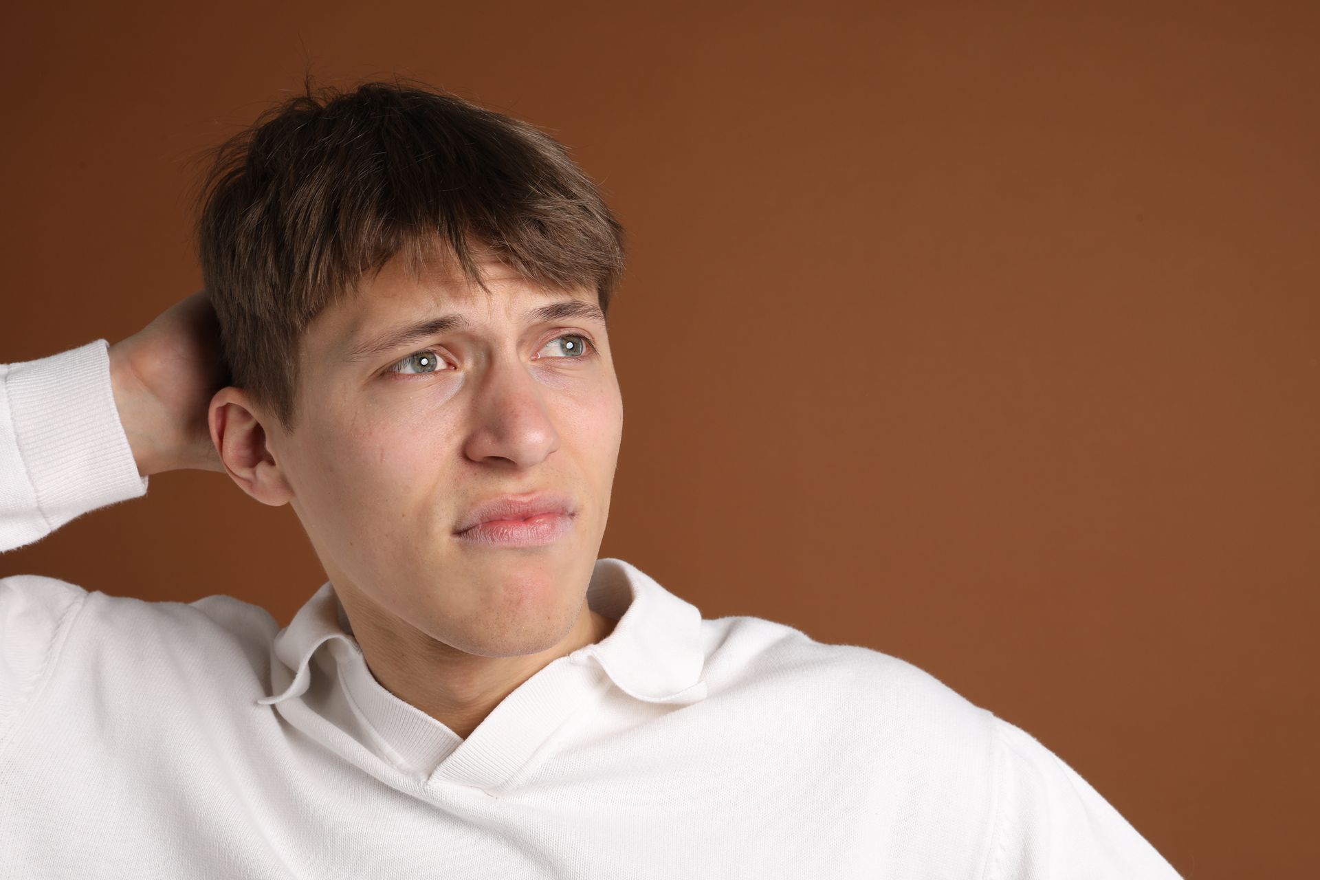 Man in white sweater, hand behind head, looking off to the side with a contemplative expression against a brown background. Man in white sweater, hand behind head, looking off to the side with a contemplative expression against a brown background.