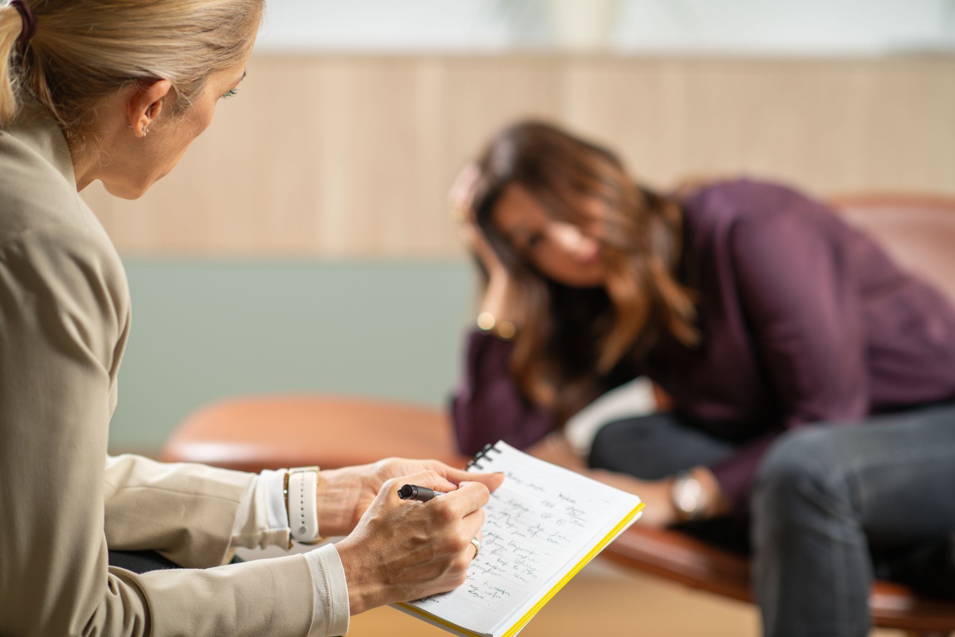 Therapist writing notes as patient looks down, resting hand on face in a consultation room. Therapist writing notes as patient looks down, resting hand on face in a consultation room.