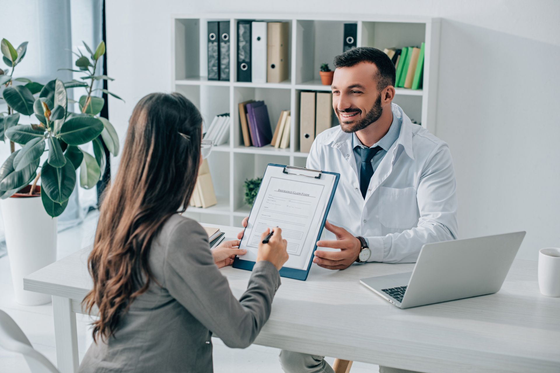 Woman in gray blazer reviews a form with a doctor in a white coat at a desk in an office. Woman in gray blazer reviews a form with a doctor in a white coat at a desk in an office.