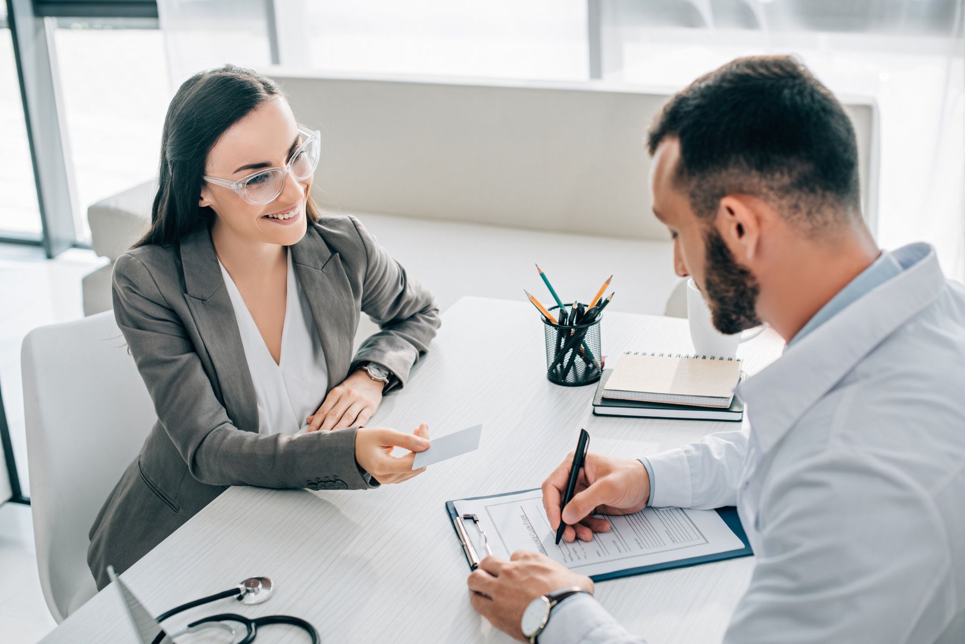 Woman handing card to doctor in a medical office. Doctor is writing on a clipboard. Woman handing card to doctor in a medical office. Doctor is writing on a clipboard.