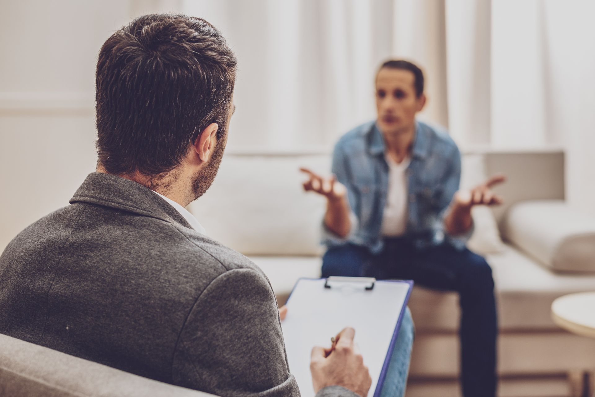 Therapist taking notes during session with a patient on a couch, gesturing with hands. Therapist taking notes during session with a patient on a couch, gesturing with hands.