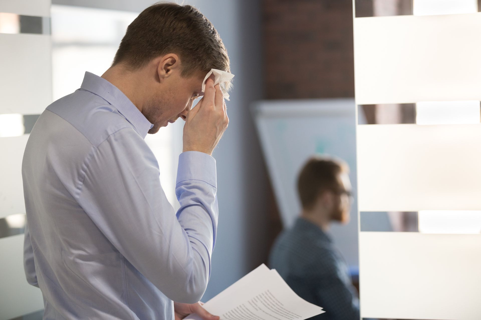 Man in light blue shirt wiping sweat from forehead, holding papers; office setting. Man in light blue shirt wiping sweat from forehead, holding papers; office setting.