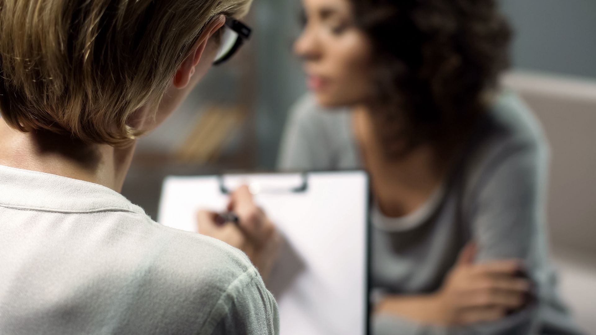 A person with glasses writes on a clipboard as another person speaks. Blurred setting suggests an office.