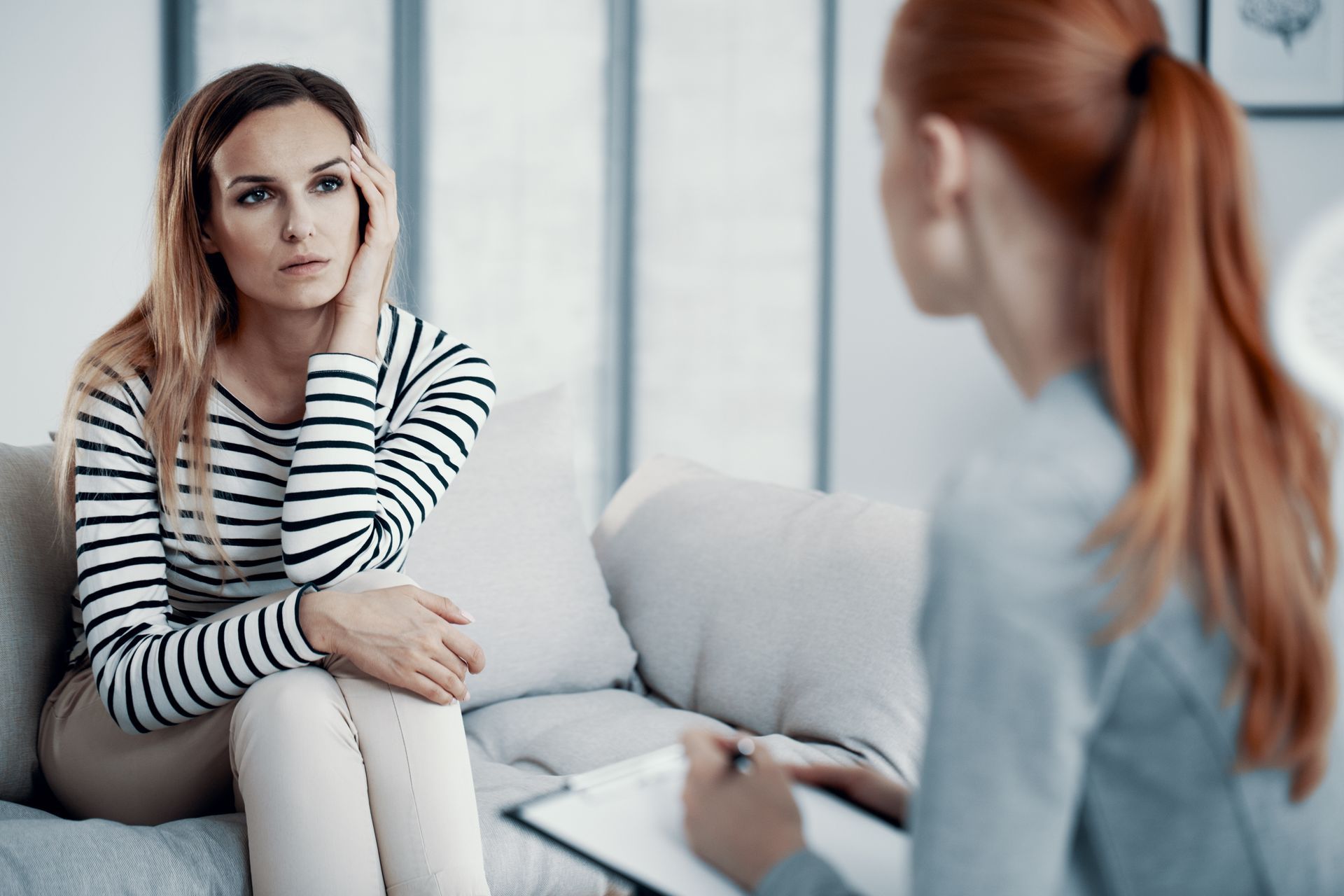 Woman on a couch with concerned expression, talking to a therapist holding a clipboard in a well-lit office. Woman on a couch with concerned expression, talking to a therapist holding a clipboard in a well-lit office.
