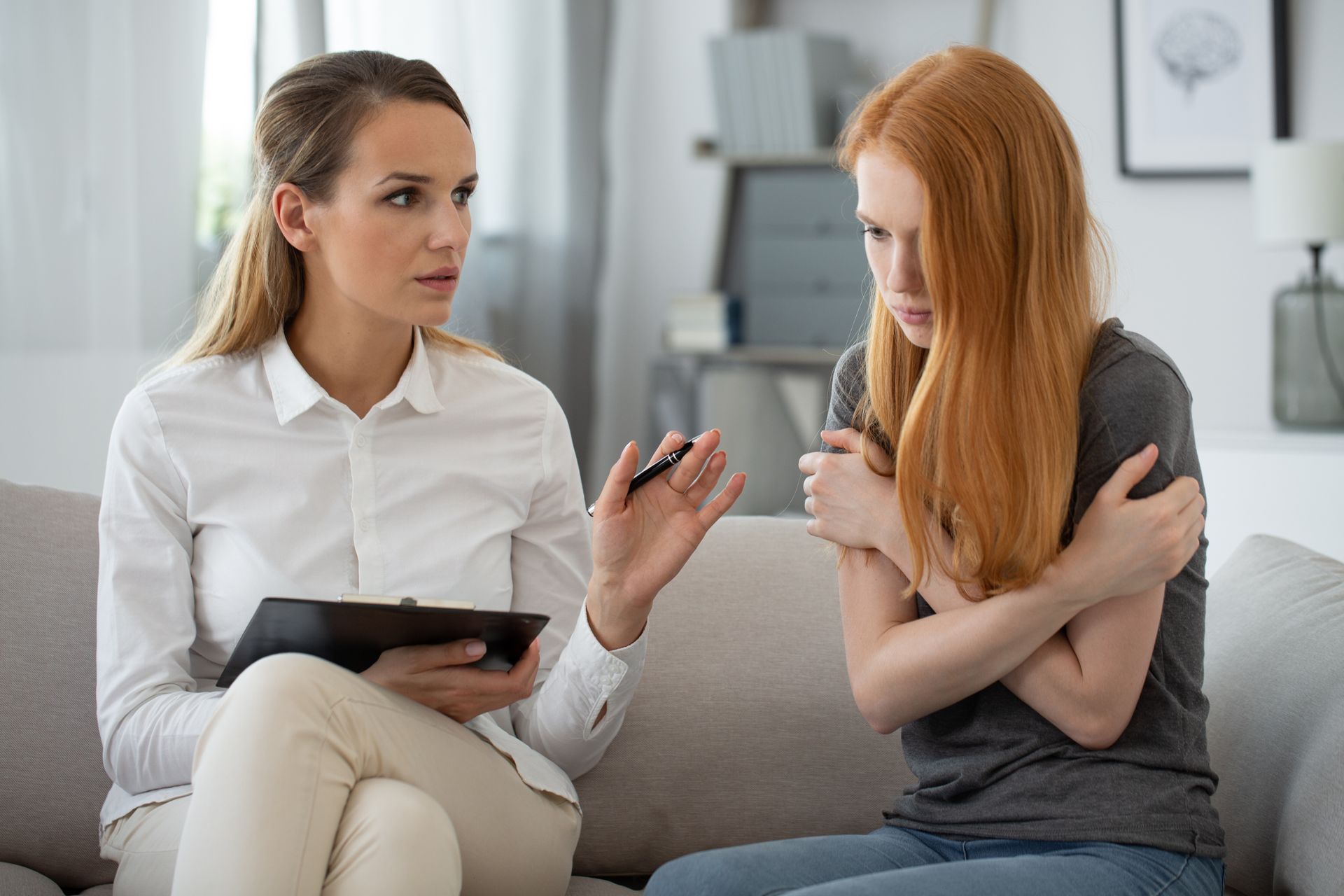 Woman in white shirt with notepad talking to a woman, arms crossed, looking concerned. Living room. Woman in white shirt with notepad talking to a woman, arms crossed, looking concerned. Living room.