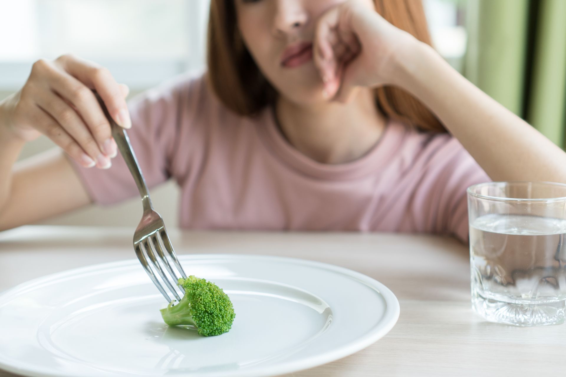 Woman with fork, broccoli on plate, looking weary, next to glass of water. Woman with fork, broccoli on plate, looking weary, next to glass of water.