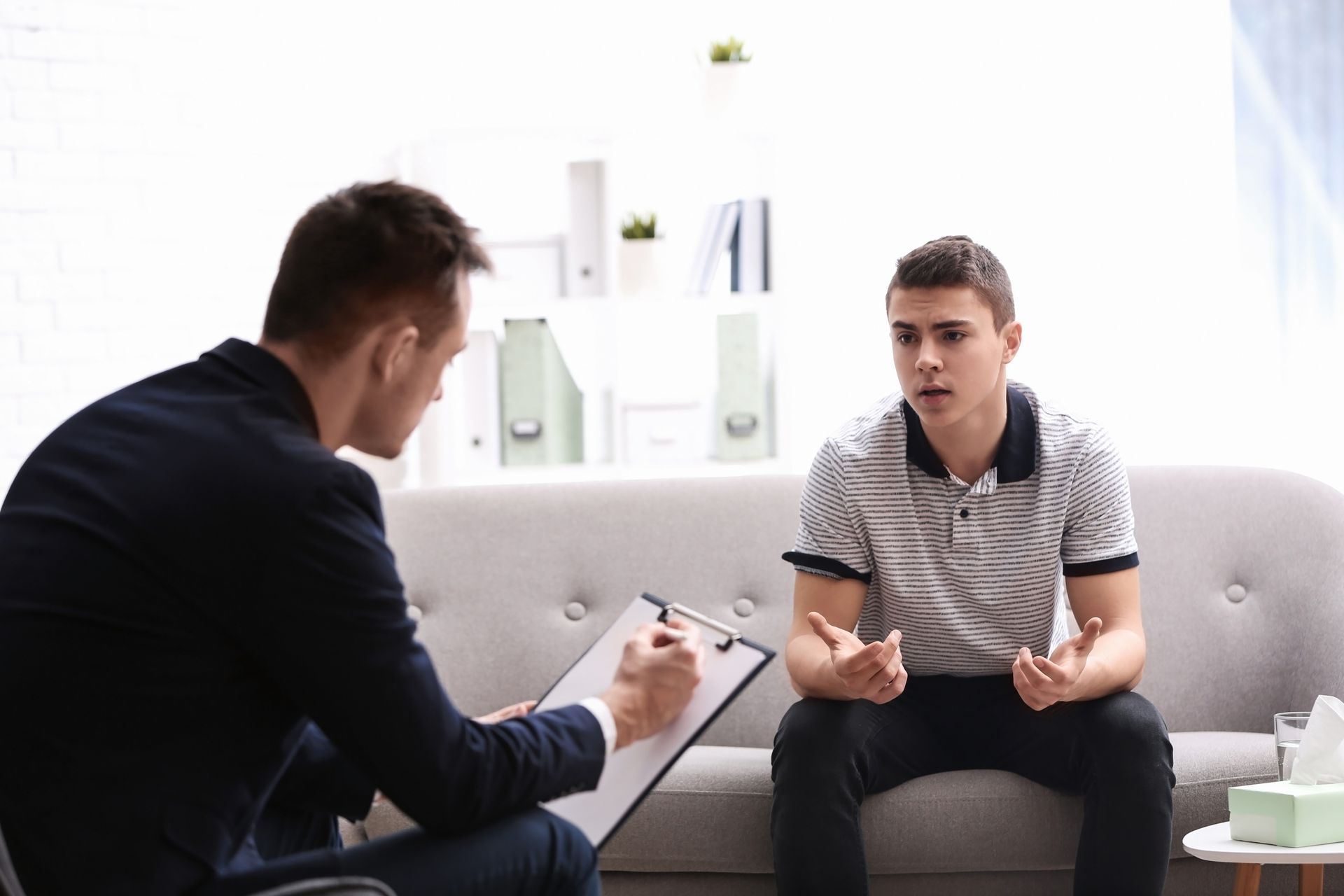 Man in suit taking notes while teenager talks on sofa in a brightly lit counseling session room. Man in suit taking notes while teenager talks on sofa in a brightly lit counseling session room.