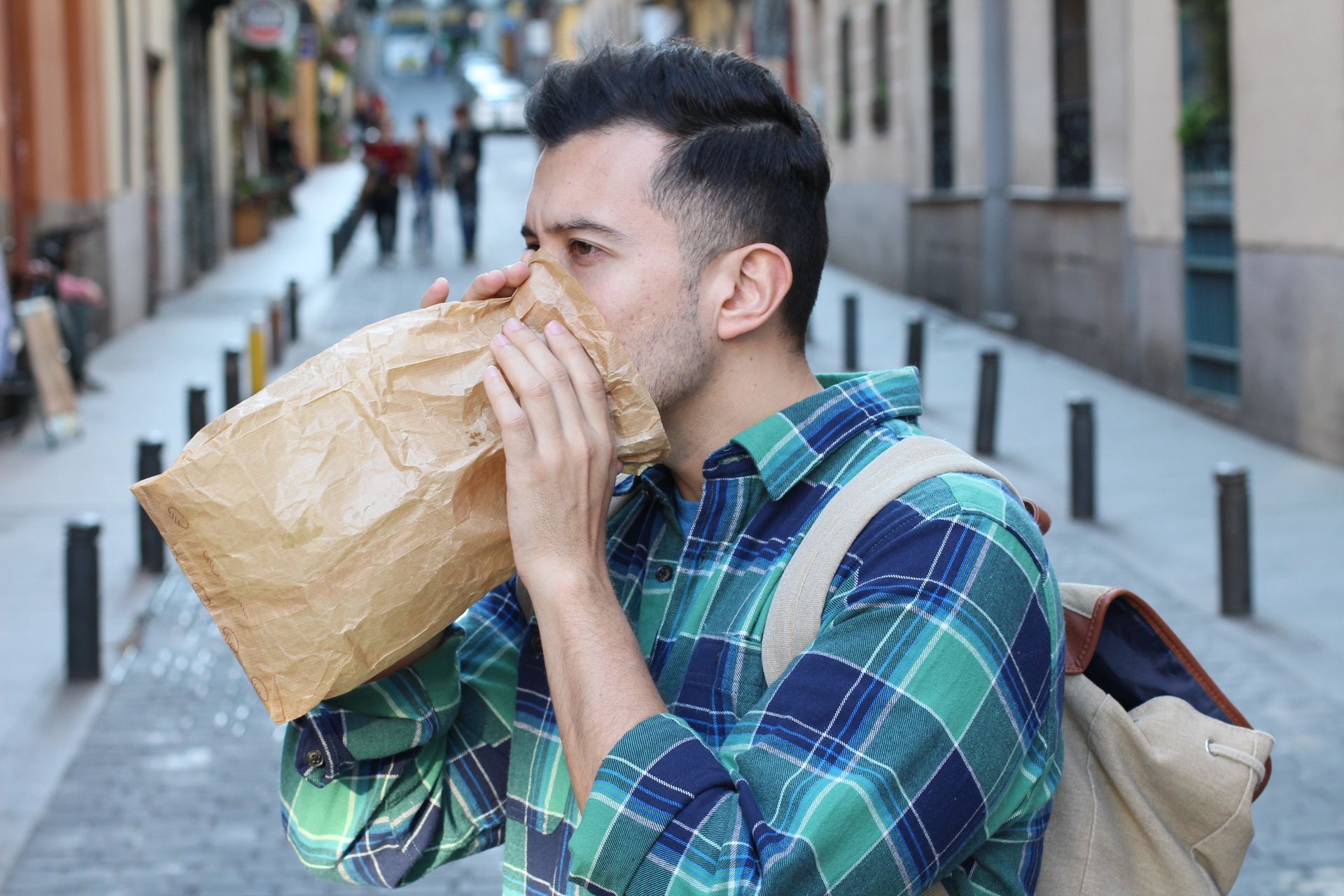 Man breathing into a brown paper bag on a city street, appearing anxious.