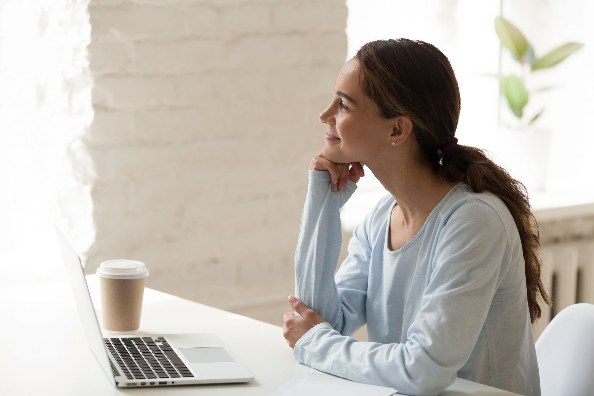 Woman at desk, looking up pensively. Laptop and coffee cup present. Light blue top, white room. Woman at desk, looking up pensively. Laptop and coffee cup present. Light blue top, white room.