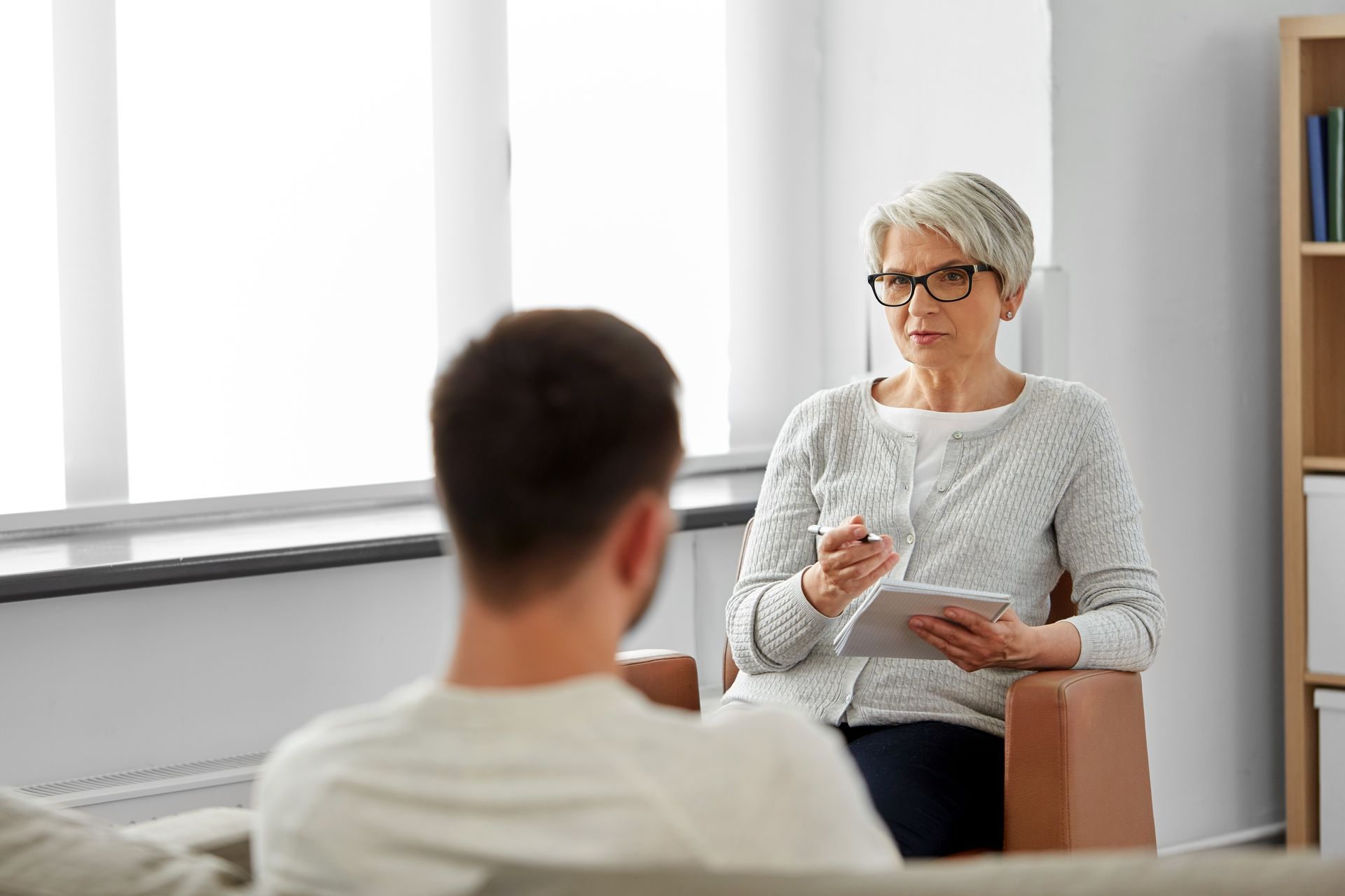 Therapist with glasses holds a tablet, speaking to a person, likely a patient, in an office setting. Therapist with glasses holds a tablet, speaking to a person, likely a patient, in an office setting.