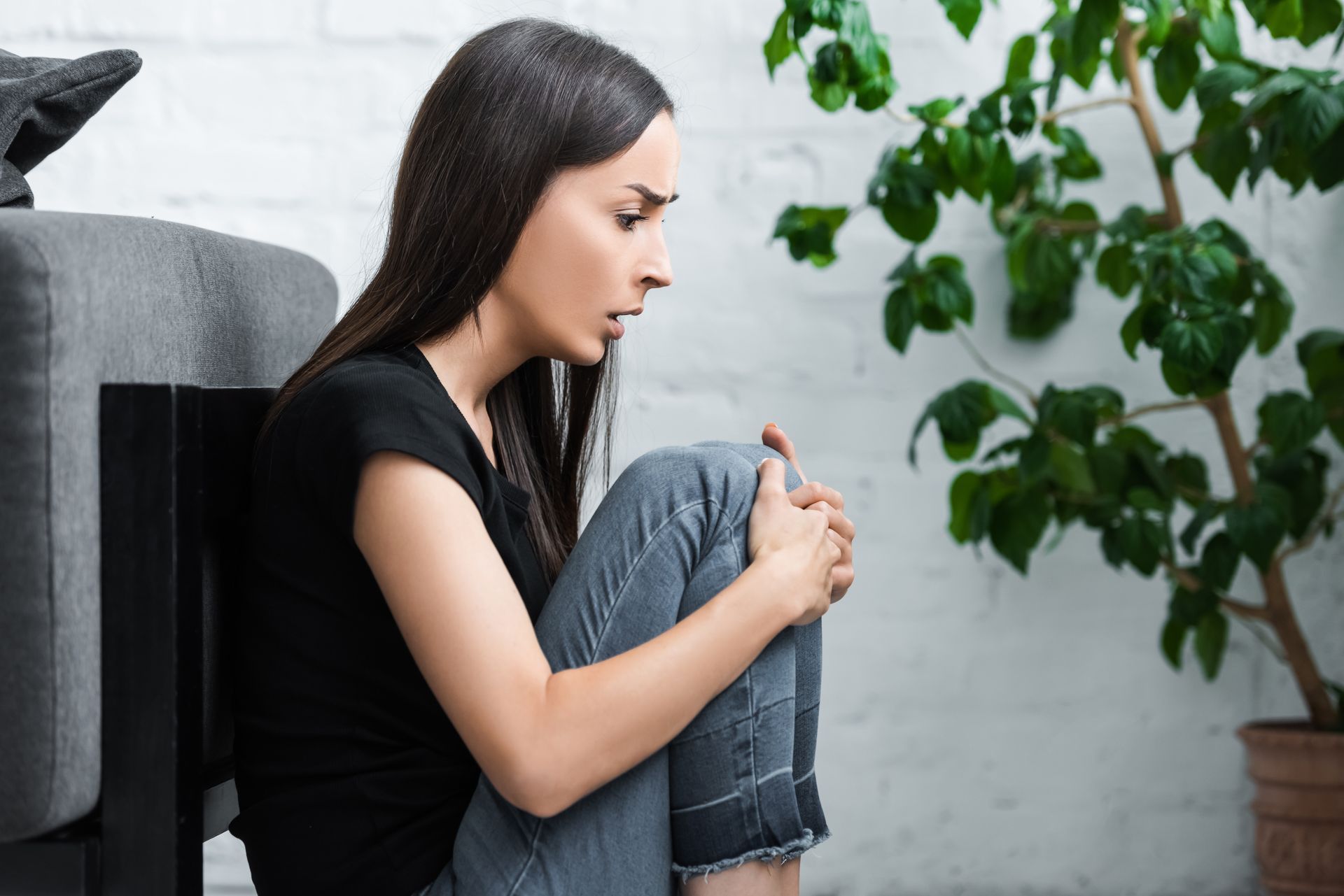Woman with knees drawn up, sitting on floor, looking distressed. Near a houseplant. Woman with knees drawn up, sitting on floor, looking distressed. Near a houseplant.