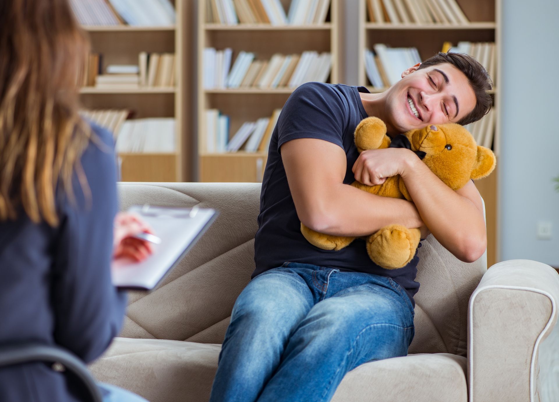 Man hugging teddy bear smiles, in therapy session with counselor, bookshelves in background. Man hugging teddy bear smiles, in therapy session with counselor, bookshelves in background.
