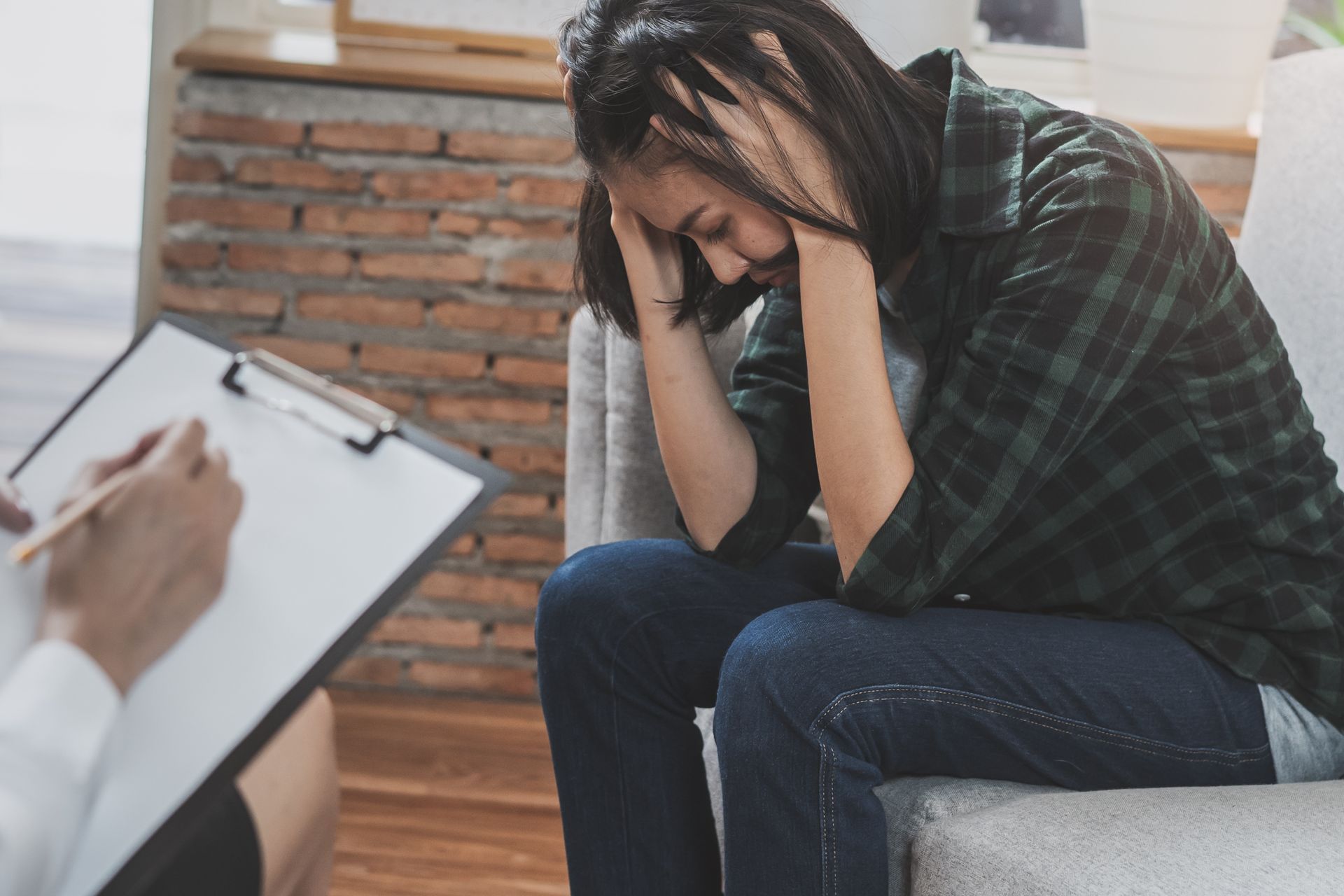 Woman with hands on head, sitting in therapy session. Therapist writing on clipboard. Woman with head in hands, seated during a counseling session. Therapist taking notes on clipboard.