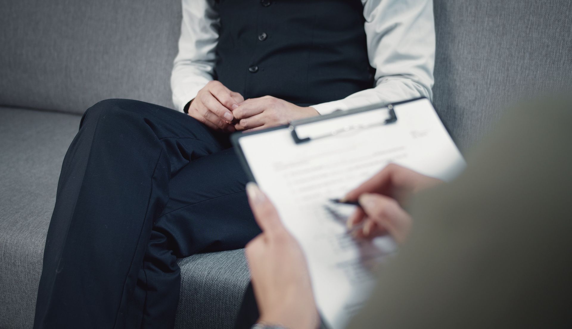 Person in vest sits on couch while another person writes on a clipboard, possibly during a therapy session.