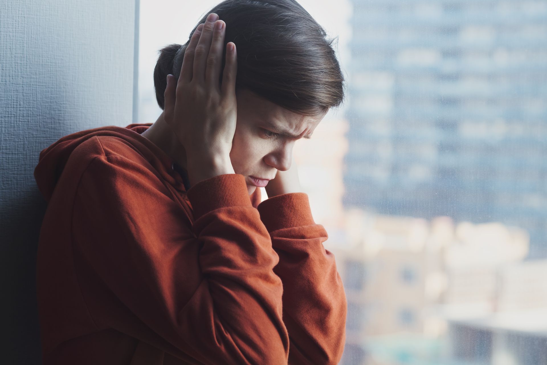 A person with hands covering ears, frowning, near a window; orange hoodie, indoor setting. A person with hands covering ears, frowning, near a window; orange hoodie, indoor setting.