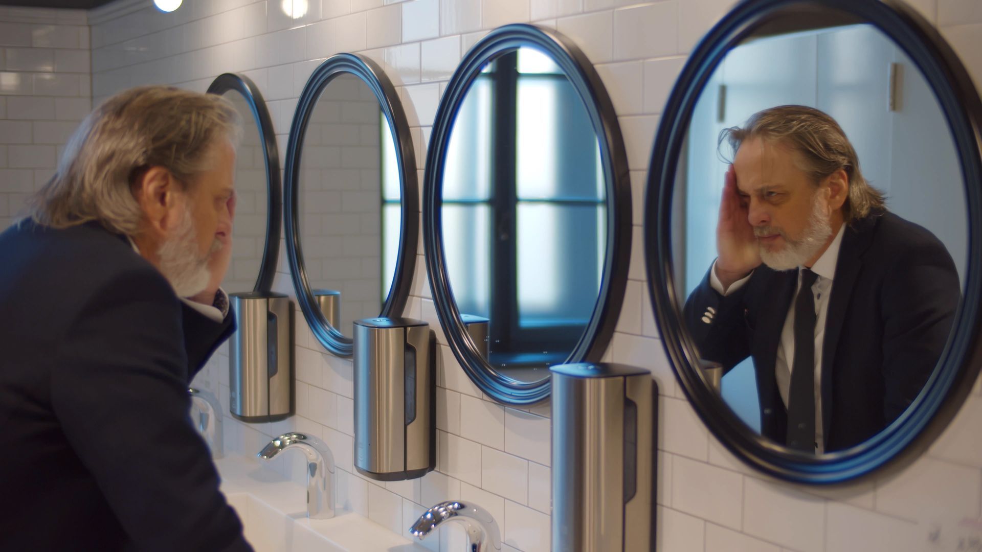 Man in suit looking into oval mirrors in a restroom, touching his head with a concerned expression. Man in suit looking into oval mirrors in a restroom, touching his head with a concerned expression.