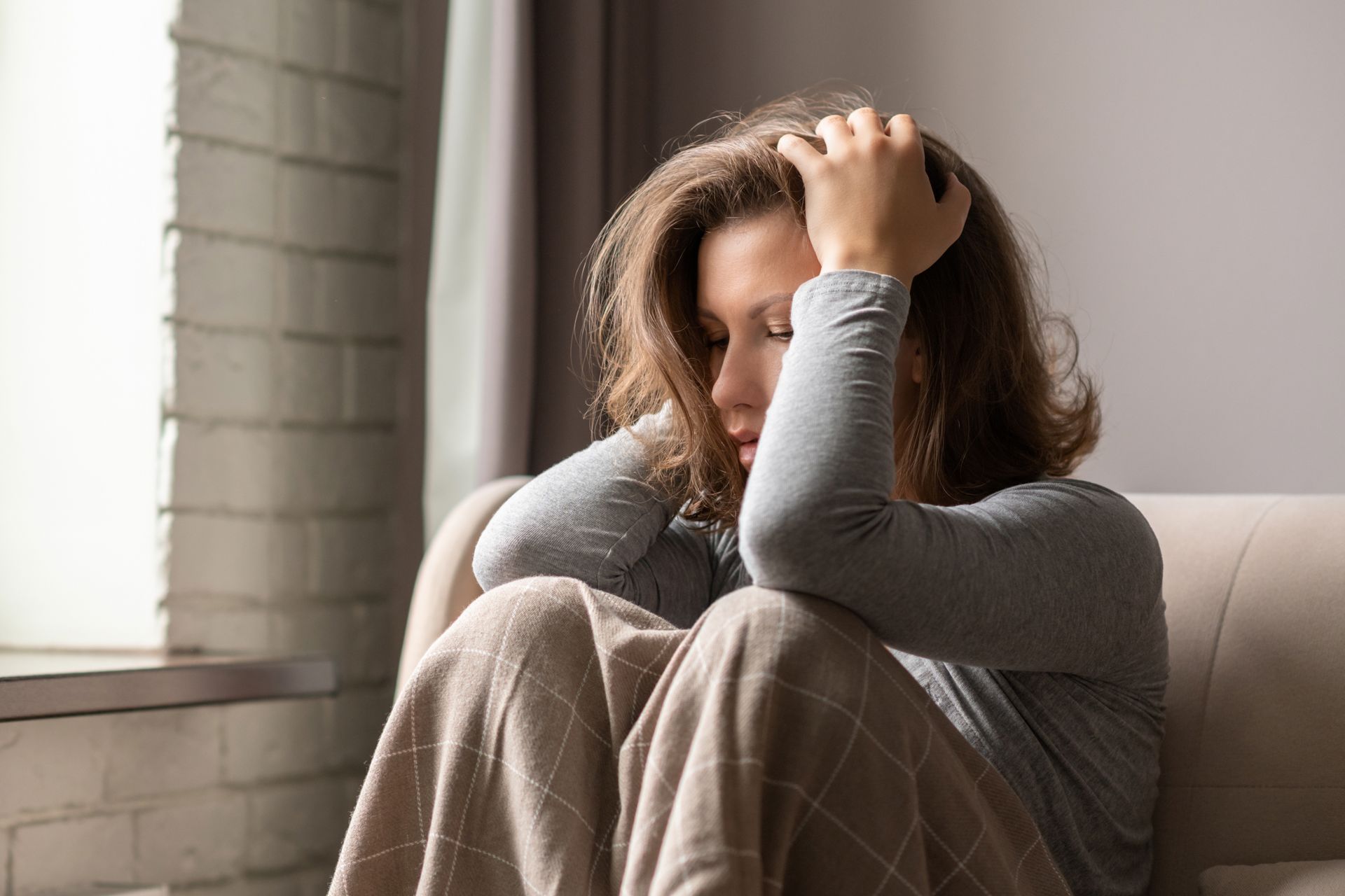 Woman sitting on a couch, holding her head with a look of distress. She is near a window. Woman sitting on a couch, holding her head with a look of distress. She is near a window.