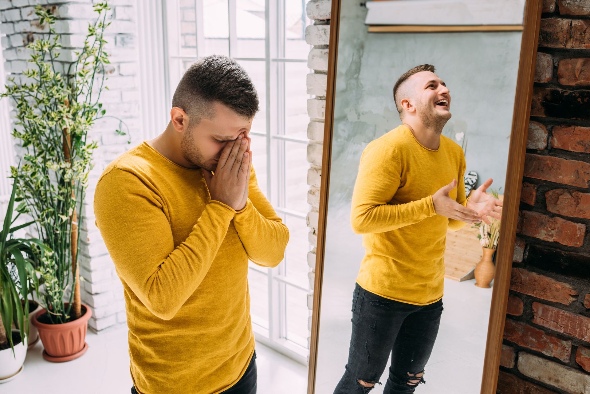 Man in yellow shirt, sad and looking at mirror reflection of himself laughing. Brick wall background.