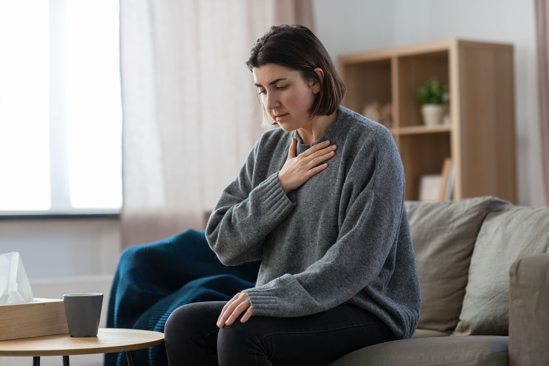 Woman clutching chest, sitting on couch, appearing distressed. Tissue box and coffee cup visible. Woman clutching chest, sitting on couch, appearing distressed. Tissue box and coffee cup visible.