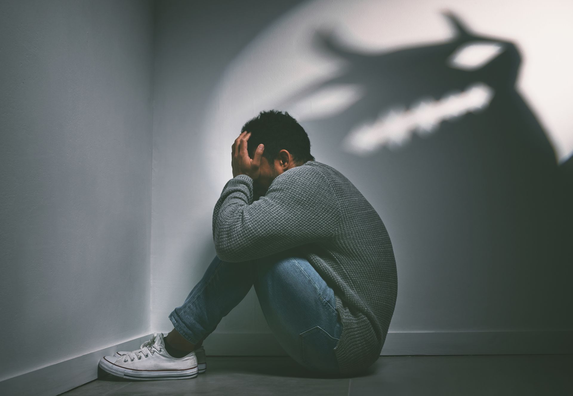 Person hunched in corner, head in hands, shadowed by ominous shape on wall.