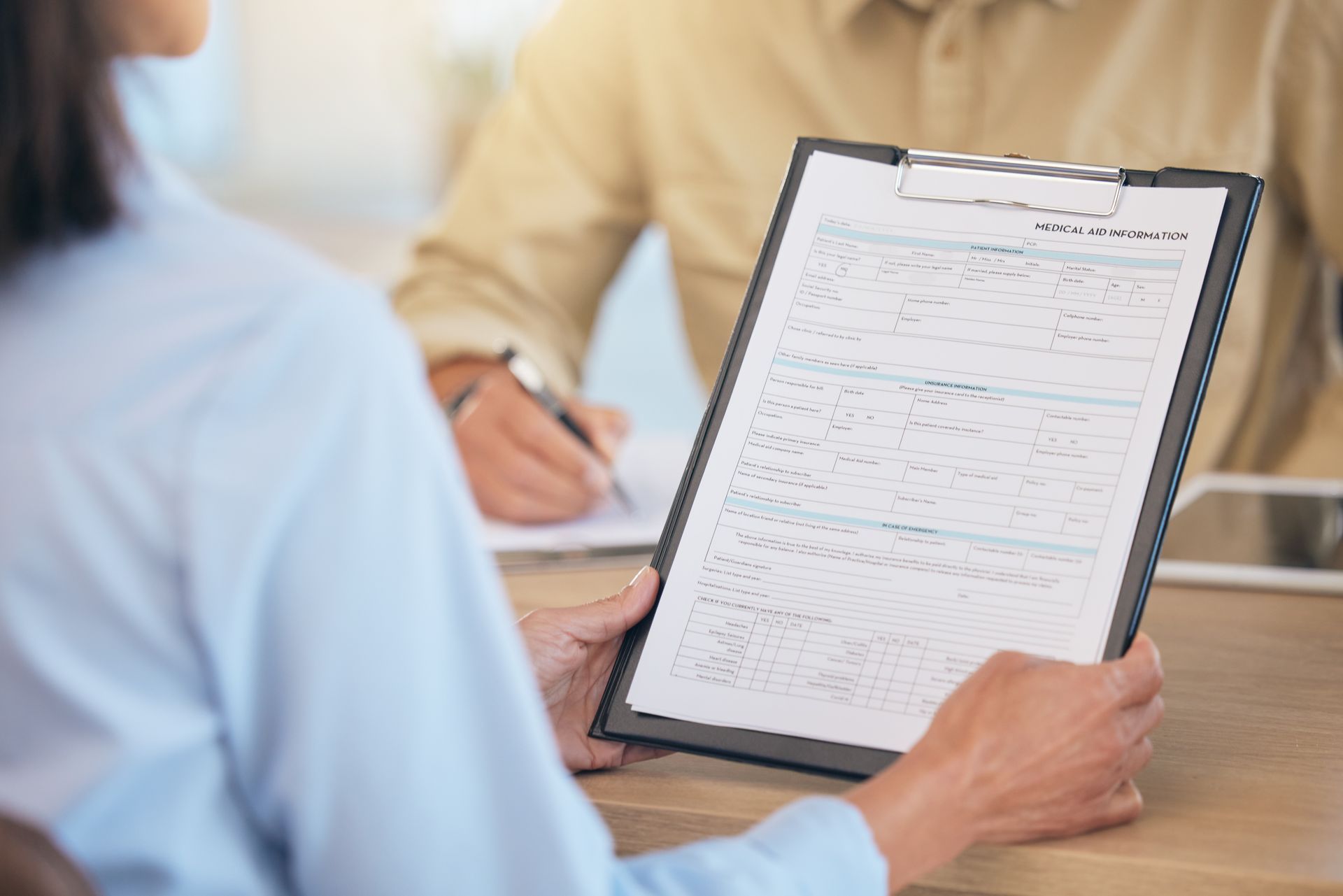 Person holding clipboard, reviewing document while another person writes at a desk. Person holding clipboard, reviewing document while another person writes at a desk.