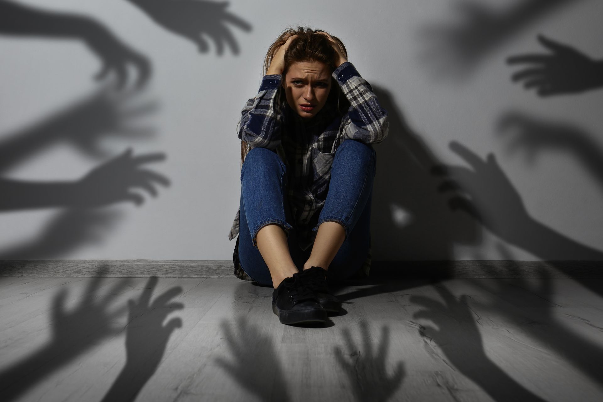 Woman sitting against a wall, looking distressed with shadows of hands reaching toward her.