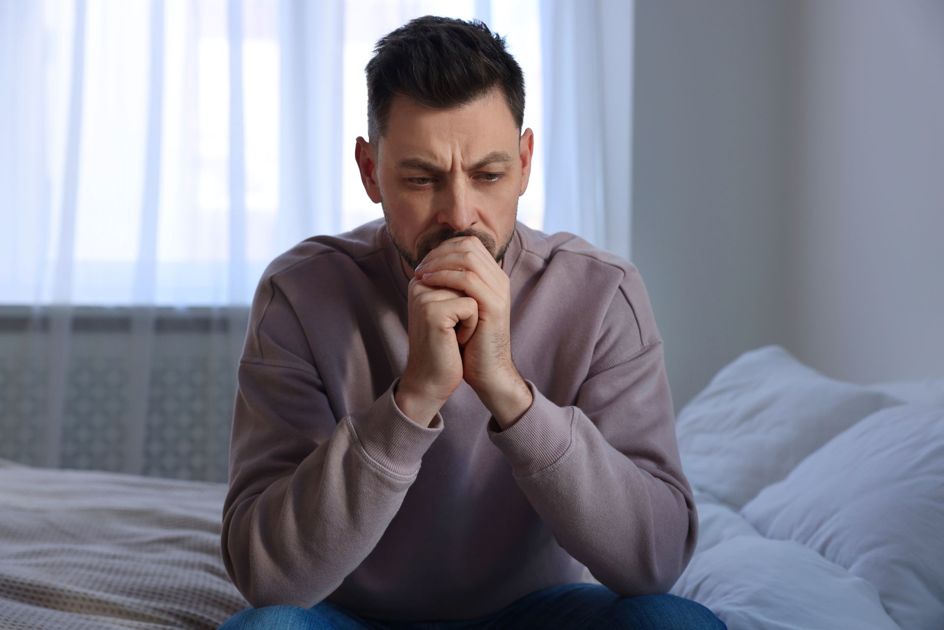 Man seated on a bed, hands clasped near chin, appearing contemplative in a bedroom with natural light. Man seated on a bed, hands clasped near chin, appearing contemplative in a bedroom with natural light.