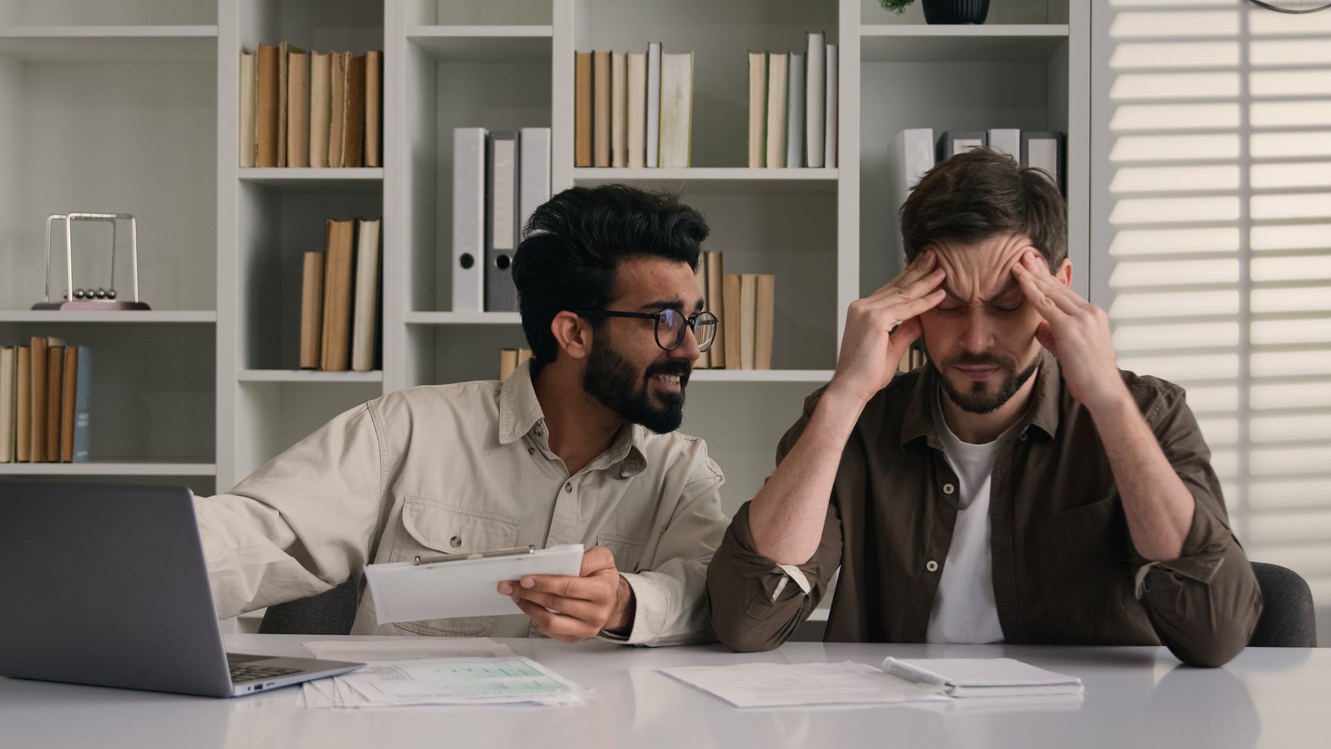 Two men at a desk, one looking stressed, hands on head; the other smiles, holding documents. Two men at a desk, one looking stressed, hands on head; the other smiles, holding documents.