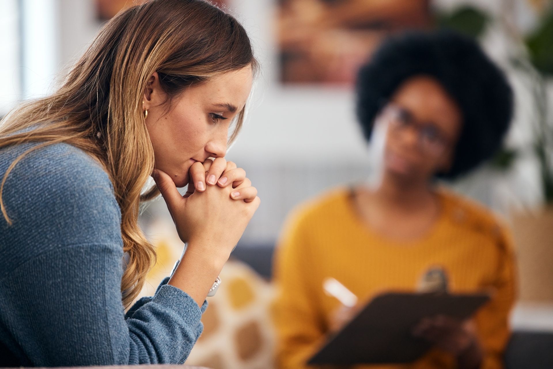 Woman in therapy session, hands clasped, looking down. Therapist in background taking notes. Woman in therapy session, hands clasped, looking down. Therapist in background taking notes.