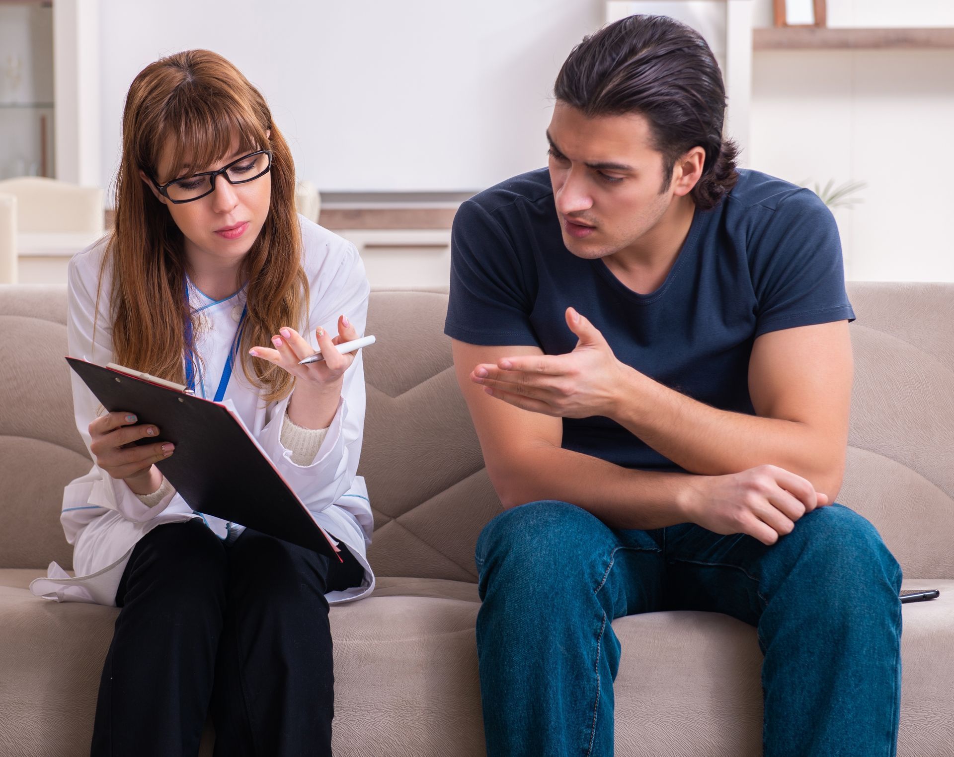Therapist with glasses takes notes as a client gestures, sitting on a sofa in an office. Therapist with glasses takes notes as a client gestures, sitting on a sofa in an office.