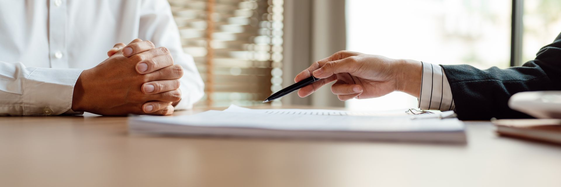 Two people at a table; one points to a document with a pen. The other person’s hands are clasped.