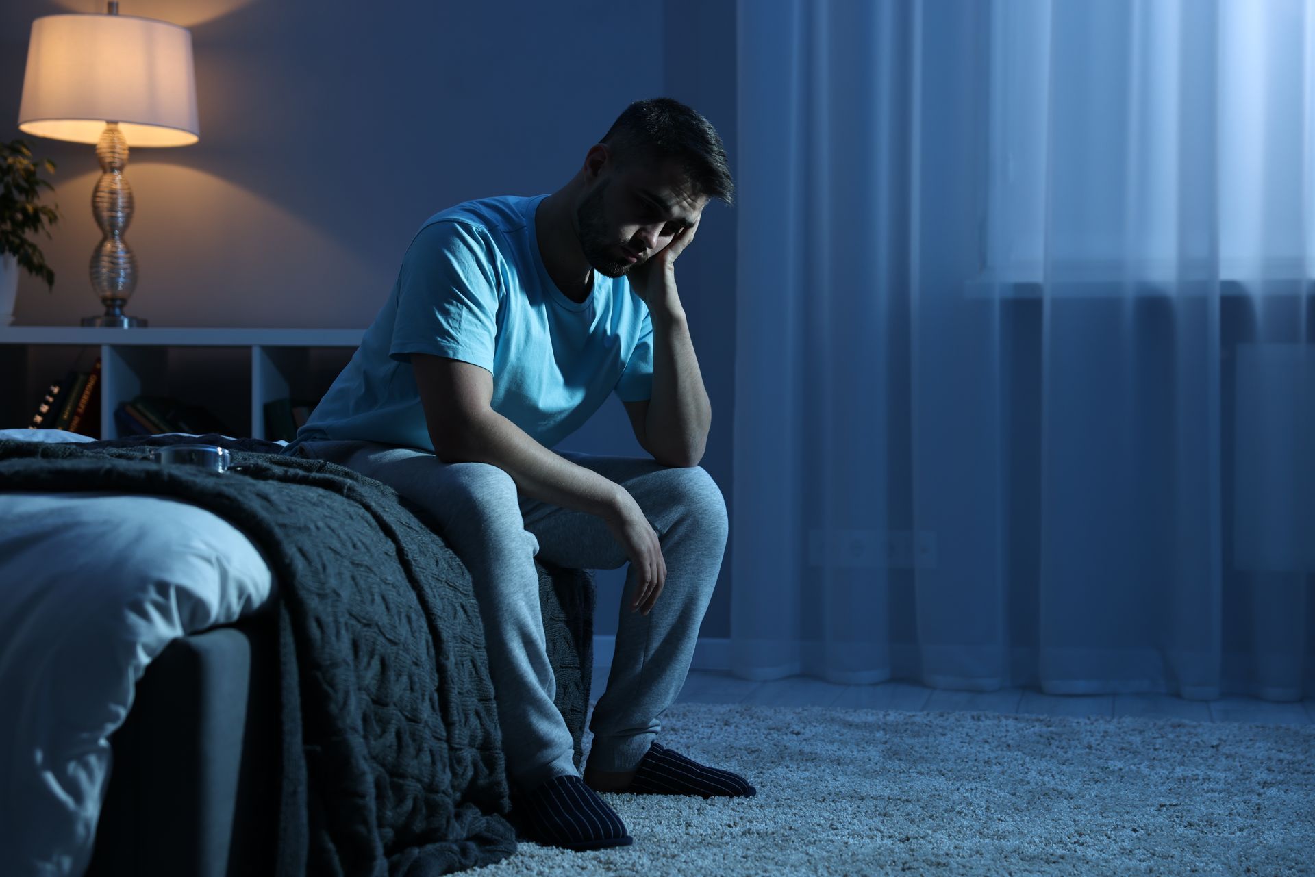 Man sitting on bed in dark room, looking down with a sad expression. Illuminated by a lamp and a window. Man sitting on bed in dark room, looking down with a sad expression. Illuminated by a lamp and a window.