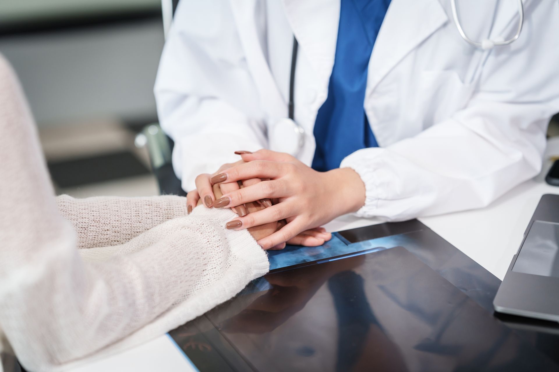 Doctor comforting patient, holding hands at a desk with an x-ray; medical office setting.