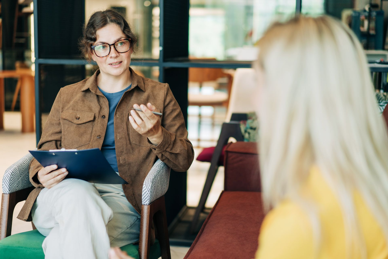 Therapist in glasses, brown jacket, blue shirt, and light pants talks to a blonde client in a bright setting. Therapist in glasses, brown jacket, blue shirt, and light pants talks to a blonde client in a bright setting.