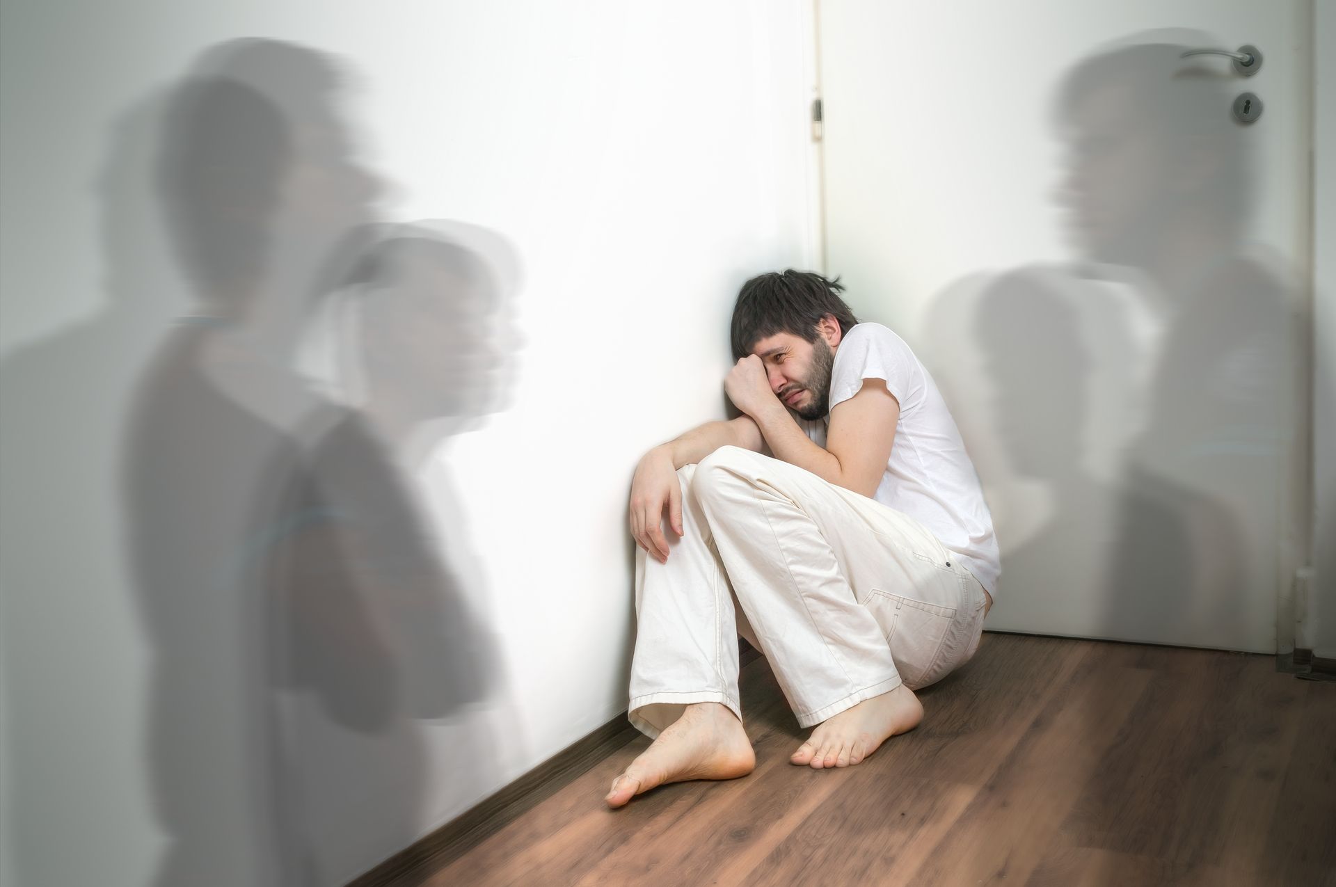 Man sits in corner, distressed. Ghostly shadows on the wall. Bare feet, white clothing, closed door. Man sits in corner, distressed. Ghostly shadows on the wall. Bare feet, white clothing, closed door.