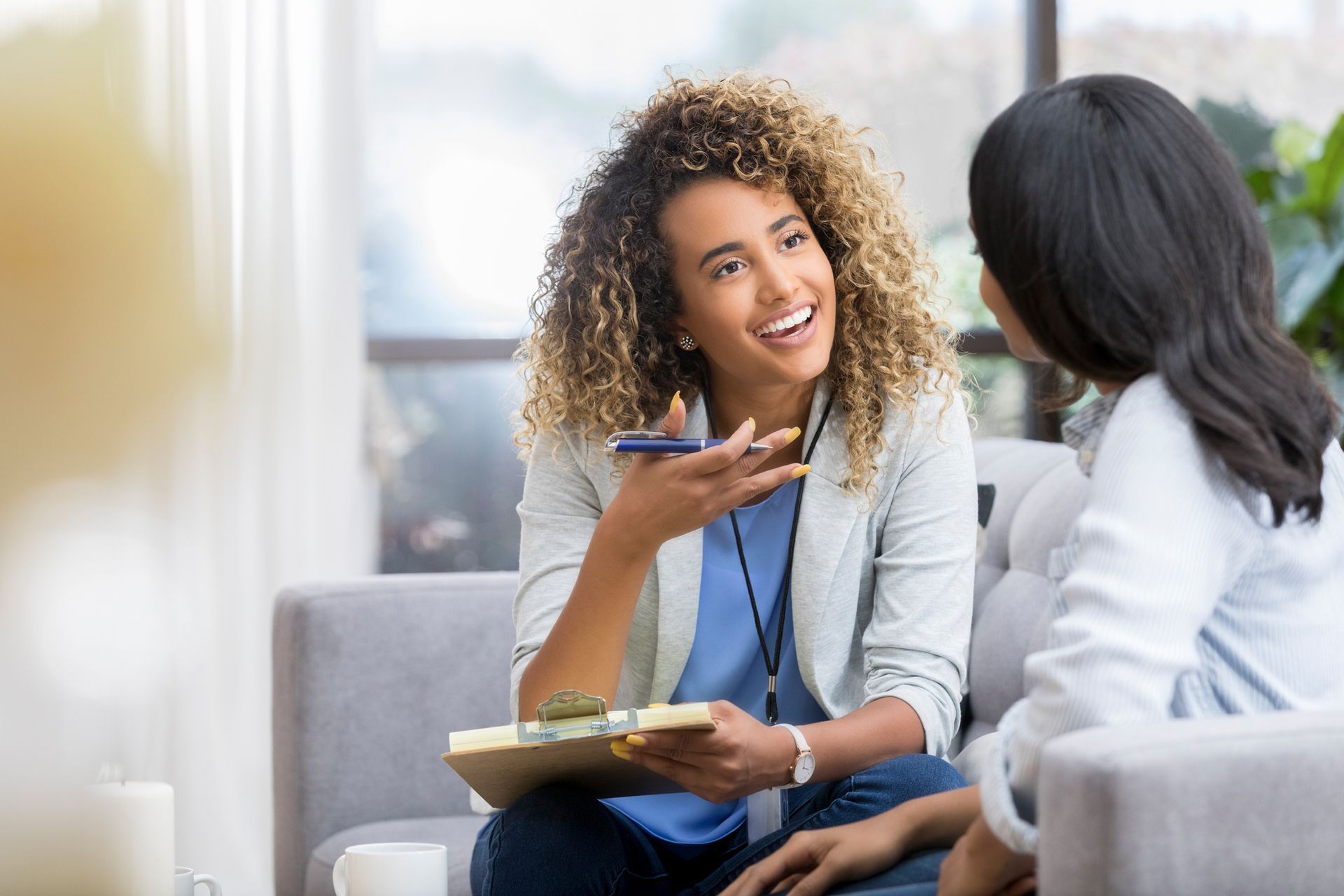 Woman with curly hair talks to another woman, holding a clipboard, indoors.