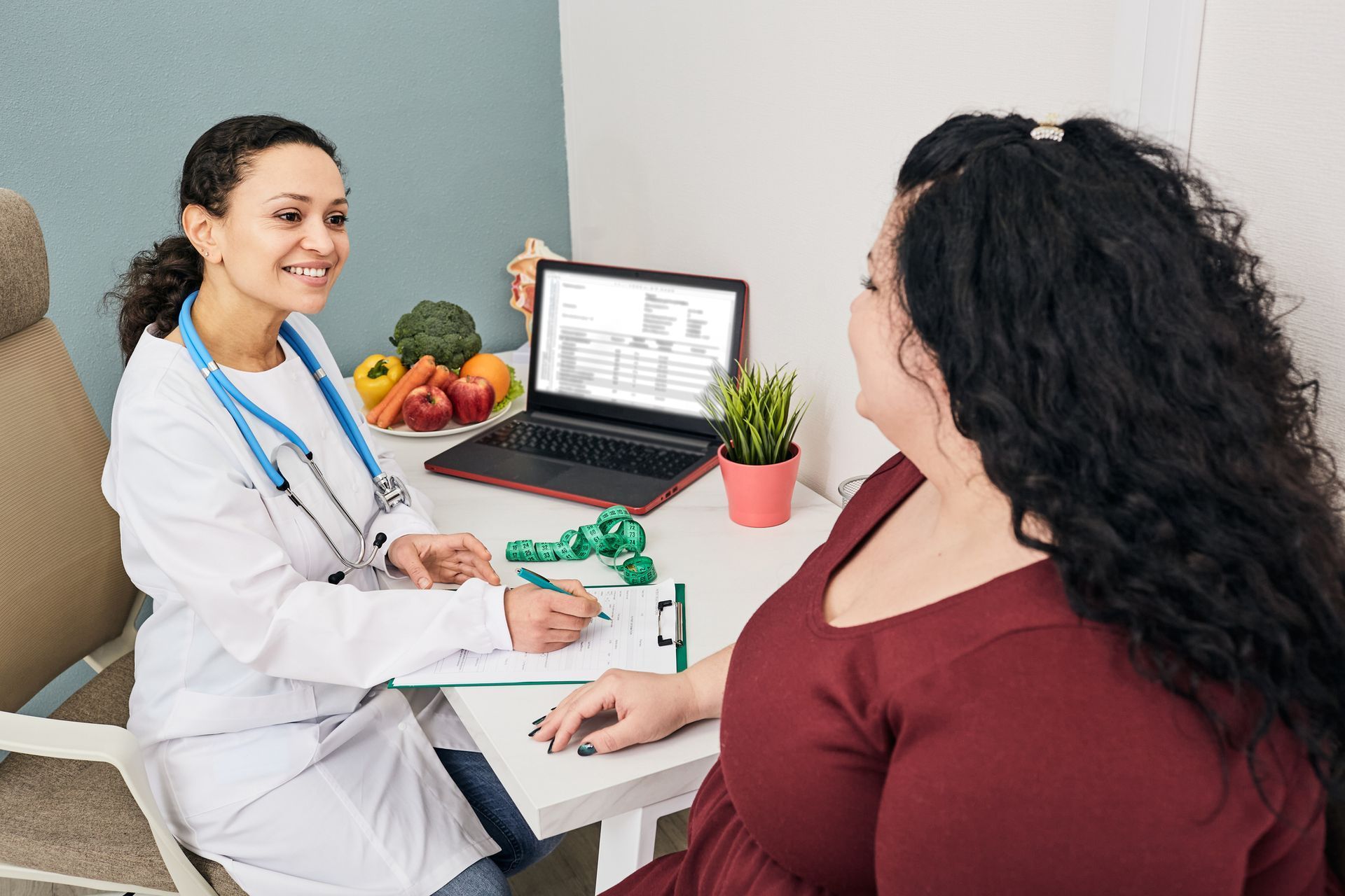 Doctor smiling at patient; examining notes. Laptop and fruits on desk. Doctor smiling at patient; examining notes. Laptop and fruits on desk.