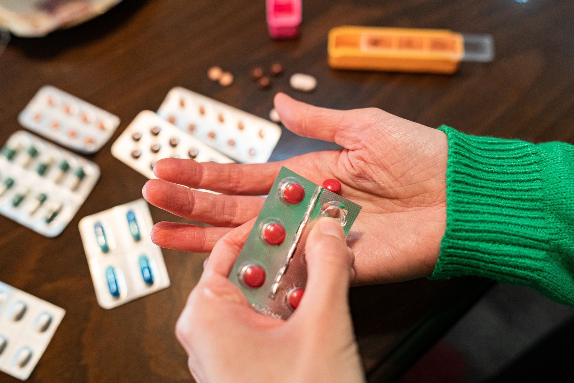 Person holding red pills in blister pack, surrounded by other medications on a table. Person holding red pills in blister pack, surrounded by other medications on a table.