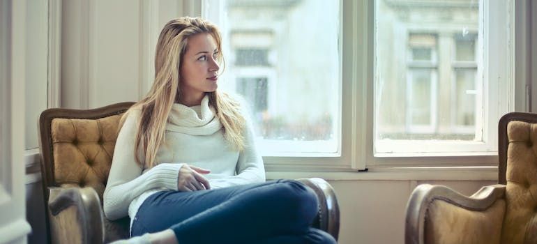 Woman in a white sweater and blue jeans sitting in a chair, looking out a window.