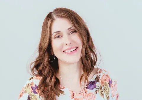Woman smiling, wearing floral top, with long wavy brown hair. Light background.